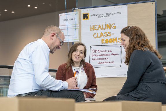 Zwei Frauen und ein Man diskutieren vor einer Stellwand mit der Aufschrift Healing Class-Rooms