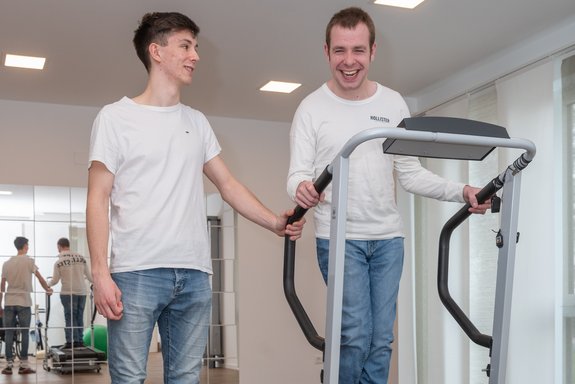 Ein junger Mann mit Handicap auf einem Sportgerät im Fitnessraum, daneben ein junger Mann ohne Handicap