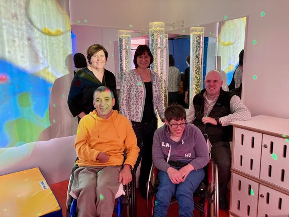 Fünf Personen, davon zwei im Rollstuhl, in einem Raum mit bunten Lichtern und beleuchteten Säulen.