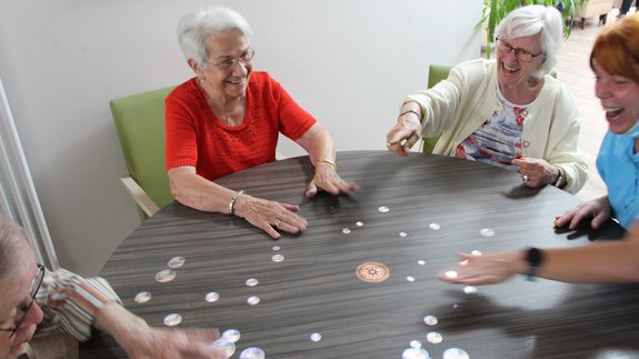 Zwei Seniorinnen und eine junge Frau sitzen an einem runden Tisch, auf den bunte Bilder projiziert werden und lachen ausgelassen.