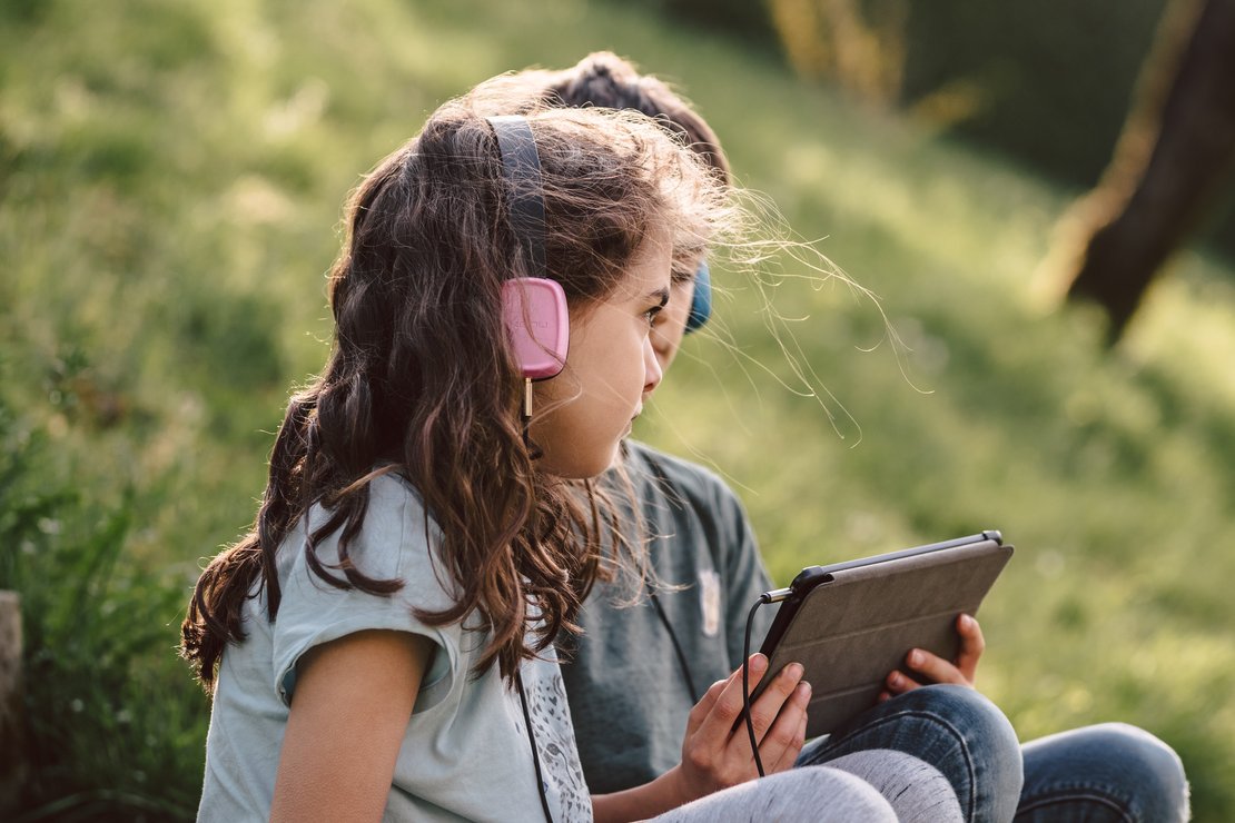 Zwei Kinder mit Kopfhörern auf dem Kopf sitzen auf einem Rasen.