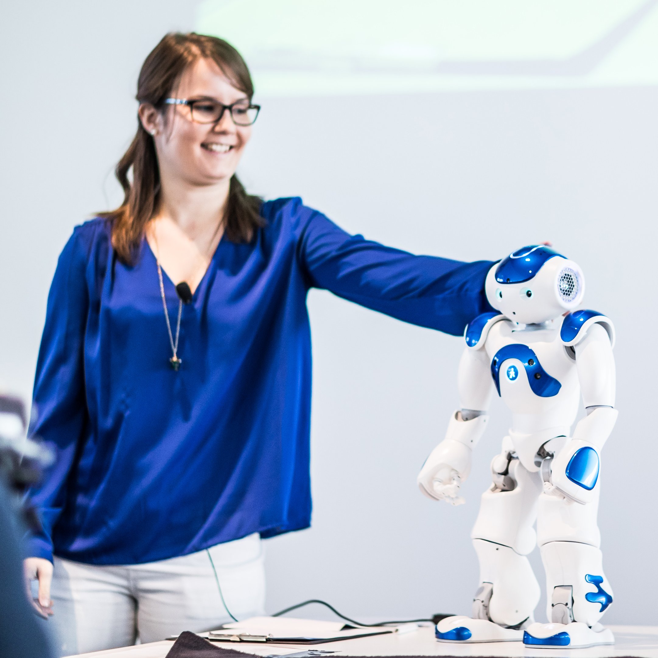Frau in blauer Bluse mit einem kleinen Roboter auf einem Tisch