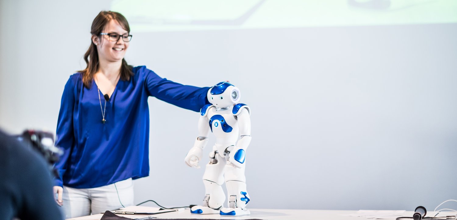 Frau in blauer Bluse mit einem kleinen Roboter auf einem Tisch