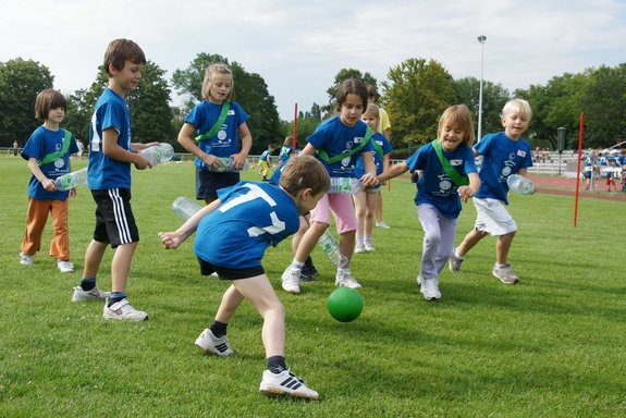 Kinder in blauen Sporttrikots spielen auf einem Rasen mit einem Ball. Sie versuchen, den Ball mit leeren Flaschen zu bewegen.