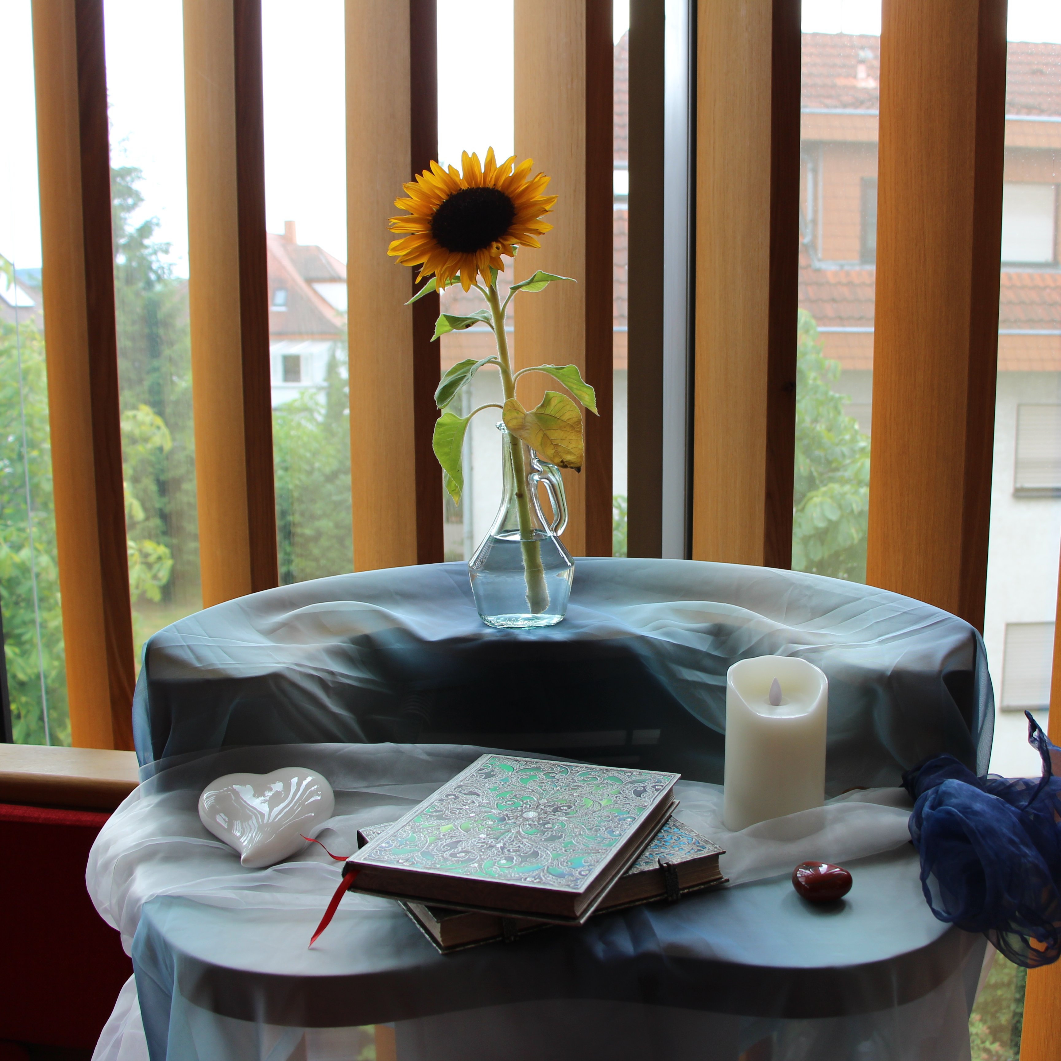 Ein runder hoher Tisch, auf dem farbenfrohe Bücher und Dekogegenstände wie Porzellanherzen, eine Kerze und eine Sonnenblume in einer Vase liegen bzw. stehen.