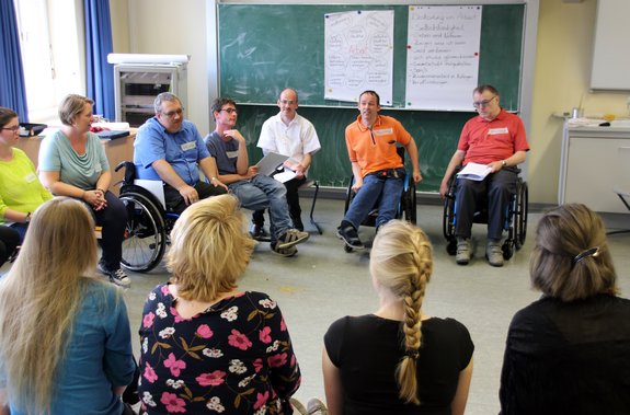 Mehrere Männer und Frauen, einige von ihnen im Rollstuhl, sitzen im Kreis. Im Hintergrund eine Schul-Tafel