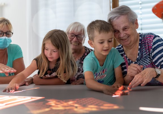 Zwei Seniorinnen haben je ein Kind auf dem Schoß, die Hände berühren bunte Bilder auf einem Tisch.