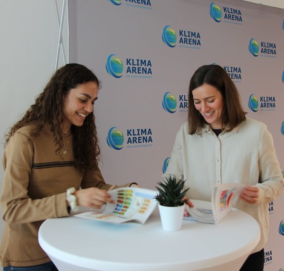 Ein Mädchen und eine junge Frau unterhalten sich lachend an einem Stehtisch, auf dem Broschüren liegen. Im Hintergrund eine Wand mit der Aufschrift Klima Arena.