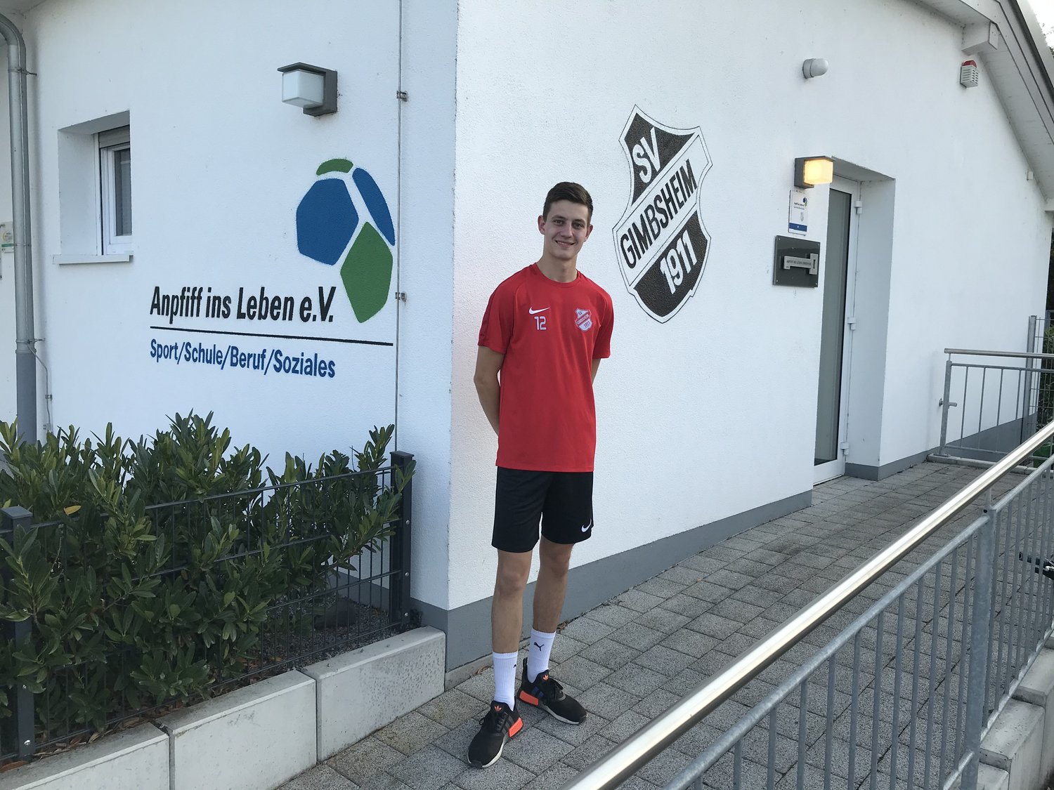 Junger Mann im Sportdress steht vor einem Clubhaus, an der Wand das Logo von Anpfiff ins Leben