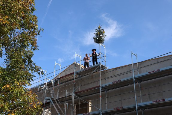 Ein Zimmermann und ein anderer Handwerker stehen auf dem Dach eines Rohbaus. Vor der Fassade steht ein Gerüst. Neben den Männern auf dem Dach ist ein baum aufgestellt.