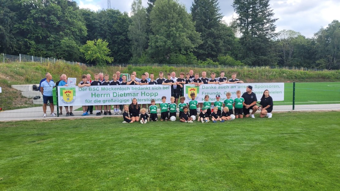 Kinder und Jugendliche in Sportkleidung an der Bande eines Fußballplatzes. An der Bande hängt ein Banner mit der Aufschrift "Der BSC Mückenloch bedankt sich bei Dietmar Hopp für die großzügige Unterstützung".