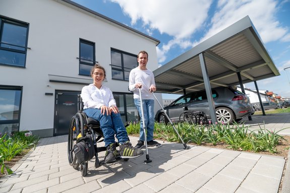 Eine Frau im Rollstuhl und ein Mann mit Gehhilfen vor einem Neubau
