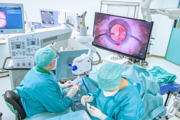 In einem Operationssaal: Der Arzt operiert und schaut dabei auf einen großen Bildschirm, auf dem ein übergroßes Auge zu sehen ist. Eine OP-Schwester bereitet Geräte vor. Im Hintergrund weitere Technik und Bildschirme.
