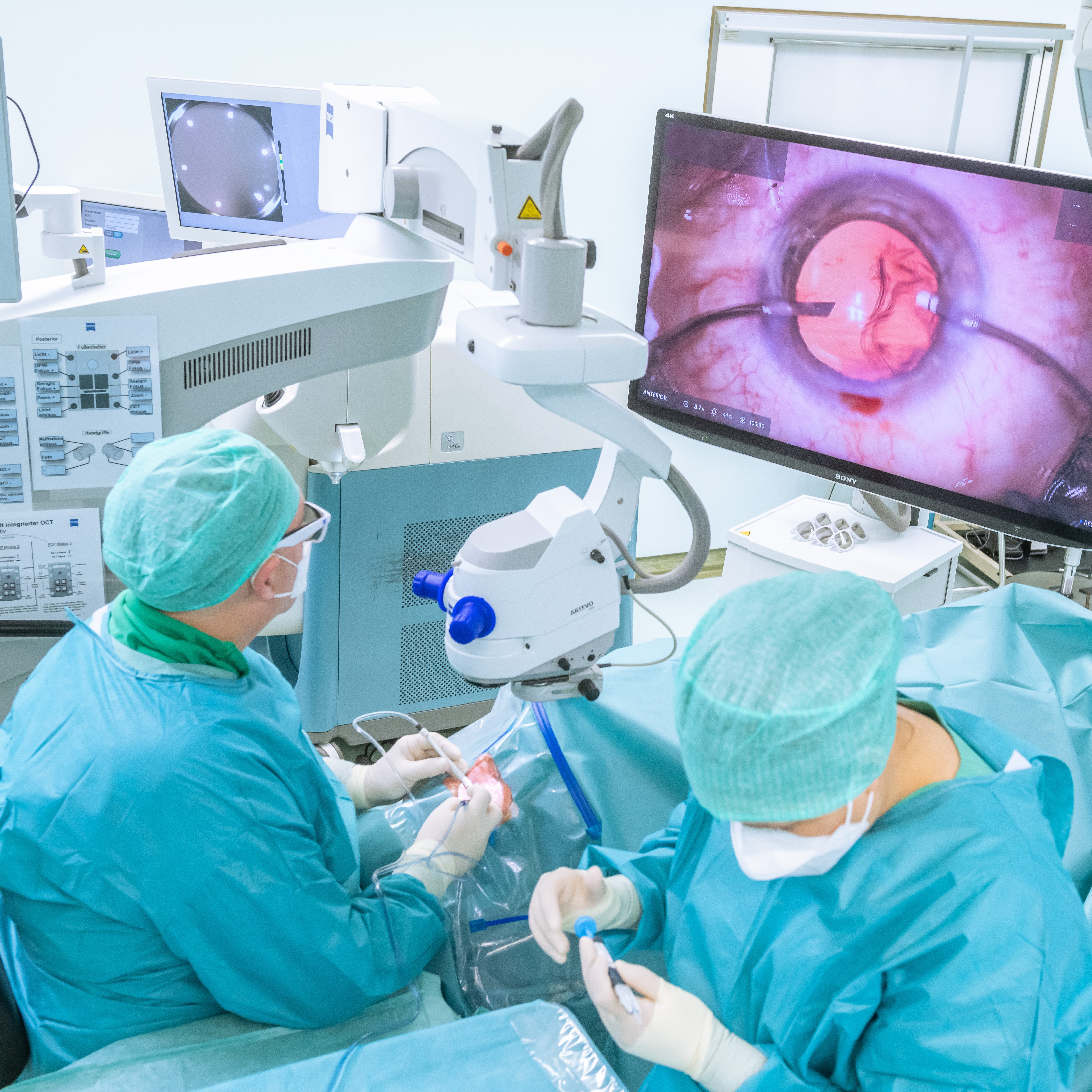Zwei Personen in OP-Kleindung in einem Operationssaal. Einer von beiden schaut auf einen Bildschirm, auf dem überdimensioniert ein Auge zu sehen ist.