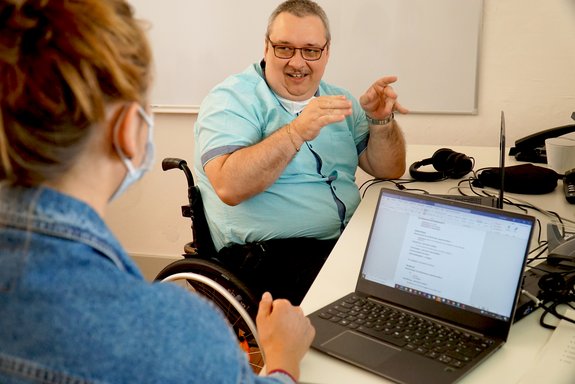 Ein Mann im Rollstuhl unterhält sich mit einer Frau, die von hinten zu sehen ist. Beide haben Laptops vor sich auf dem Tisch.