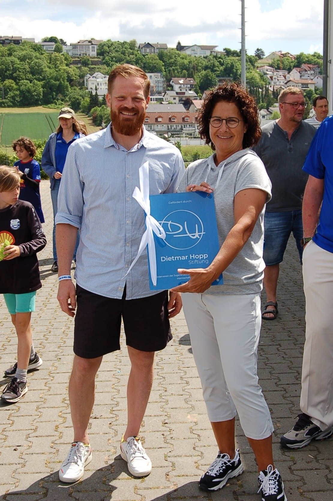 Ein Mann und eine Frau halten ein blaues Schild in die Kamera. Aufschrift: "Gefördert durch die Dietmar Hopp Stiftung"."
