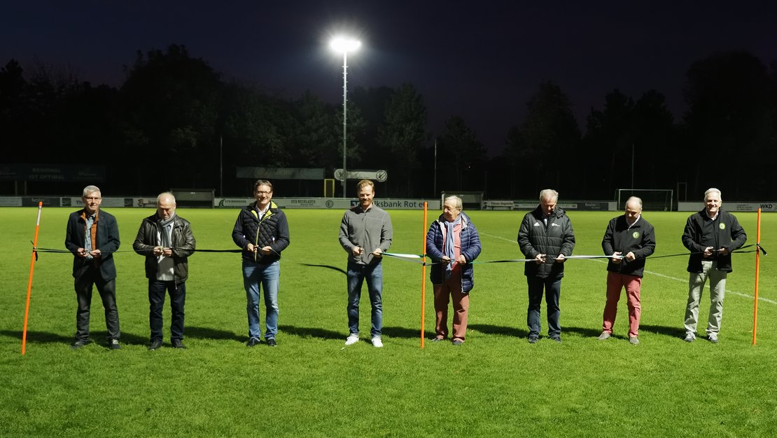 Acht Männer nebeneinander abends auf einem Fußballfeld unter Flutlicht schneiden ein Band durch.