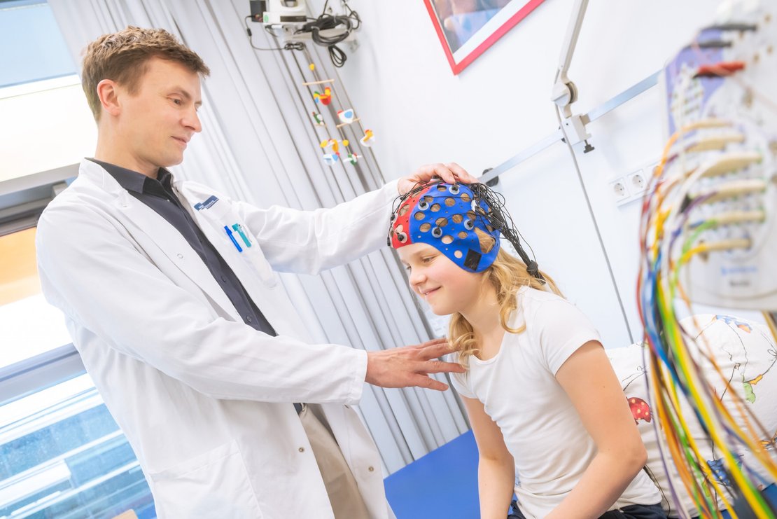 Ein Kind trägt eine Kappe mit zahlreichen Kabeln auf dem Kopf, ein Arzt steht davor und prüft den Sitz der Kappe.